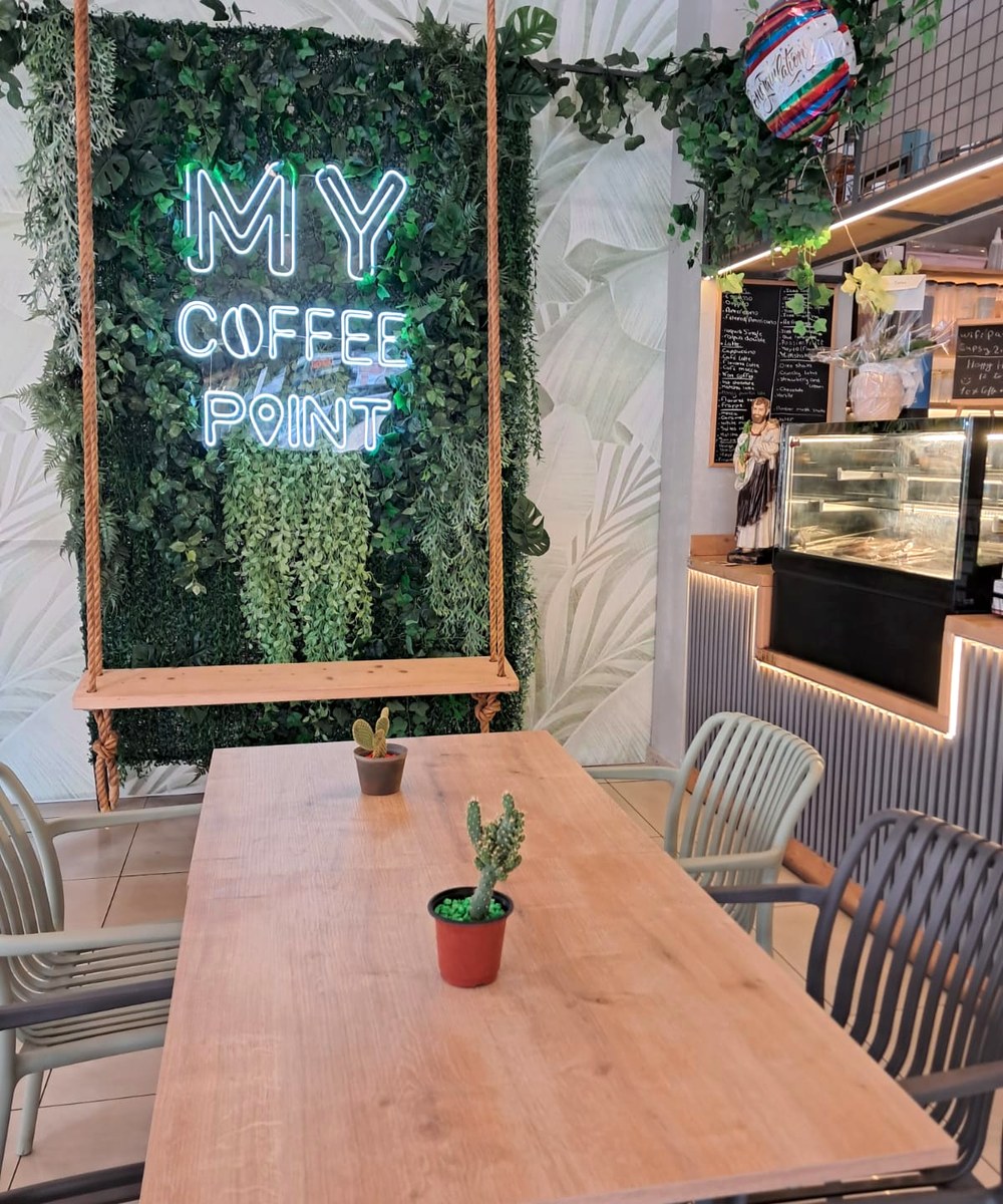 Neon 'My Coffee Point' sign above a wooden swing on a green plant wall